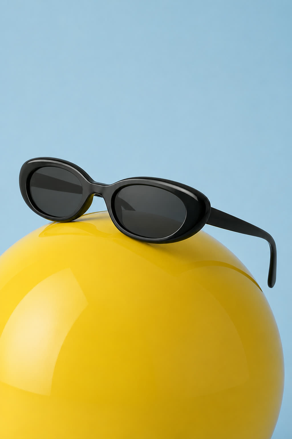 Black acetate sunglasses produced resting on a yellow sphere against a blue ground