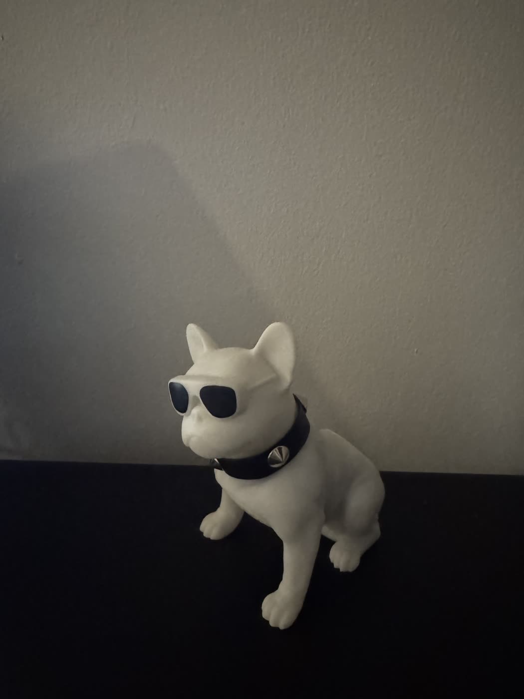 Phone photograph of a white bulldog figurine wearing sunglasses and a spiked collar, on a dark table against a plain wall
