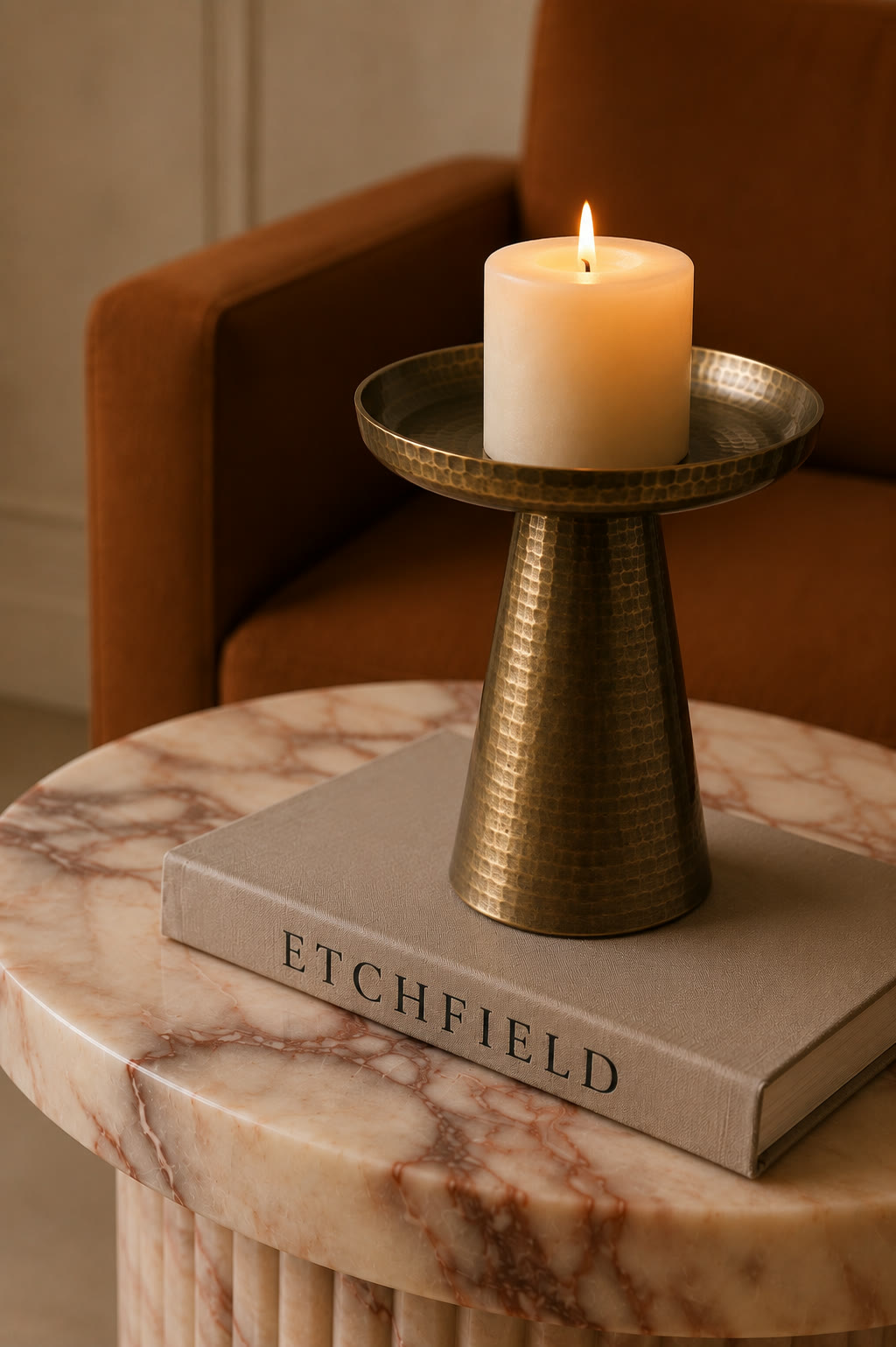 A hammered brass candle holder with a lit pillar candle, on a book on a fluted marble side table beside a rust velvet chair