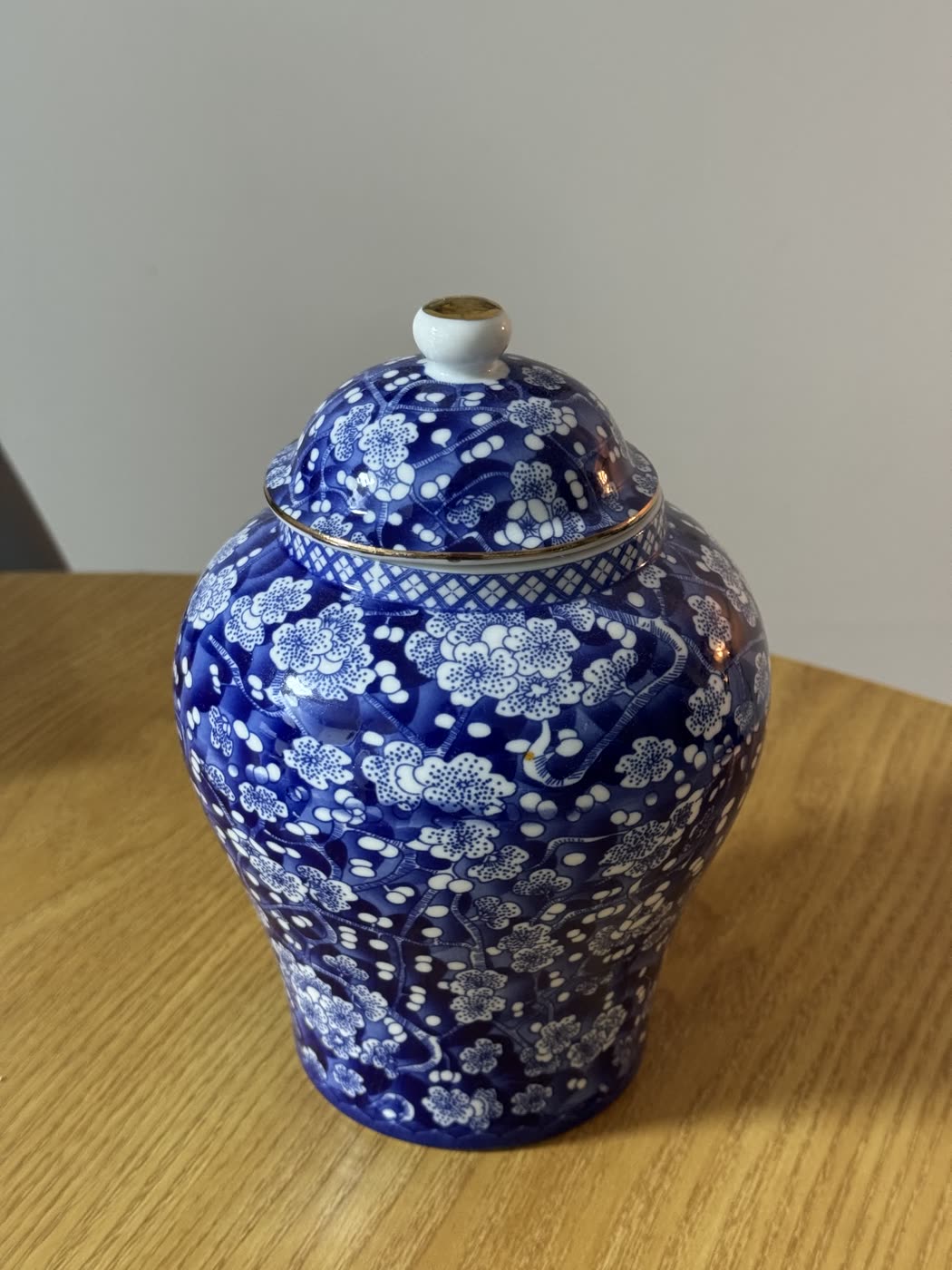 Phone photograph of a blue and white ginger jar with a gilt-rimmed lid, standing on a wooden table against a plain wall