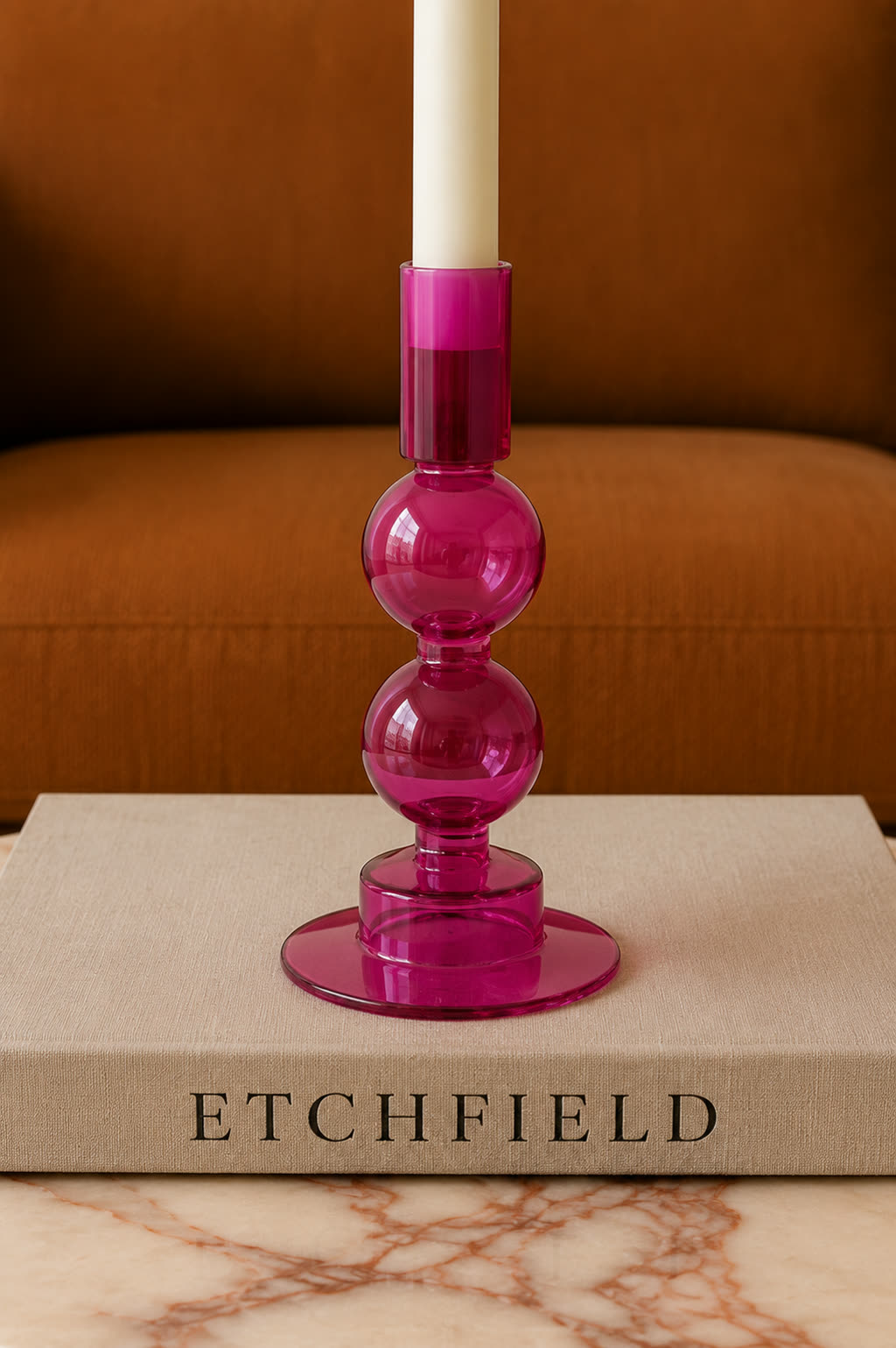 A magenta glass candle holder with a pillar candle, on a book beside a marble ledge