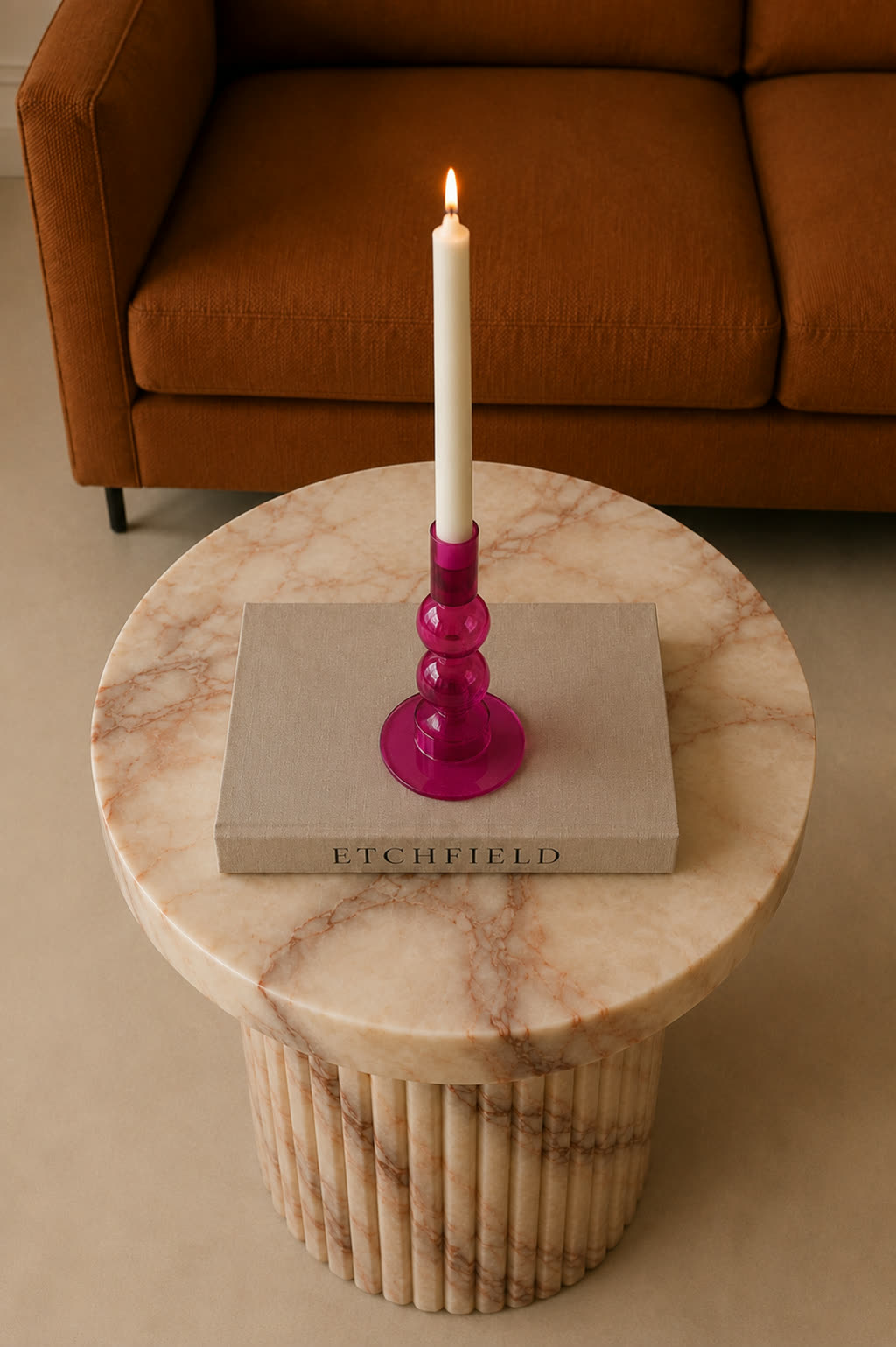 The same magenta glass candle holder with the candle lit, on a fluted marble side table