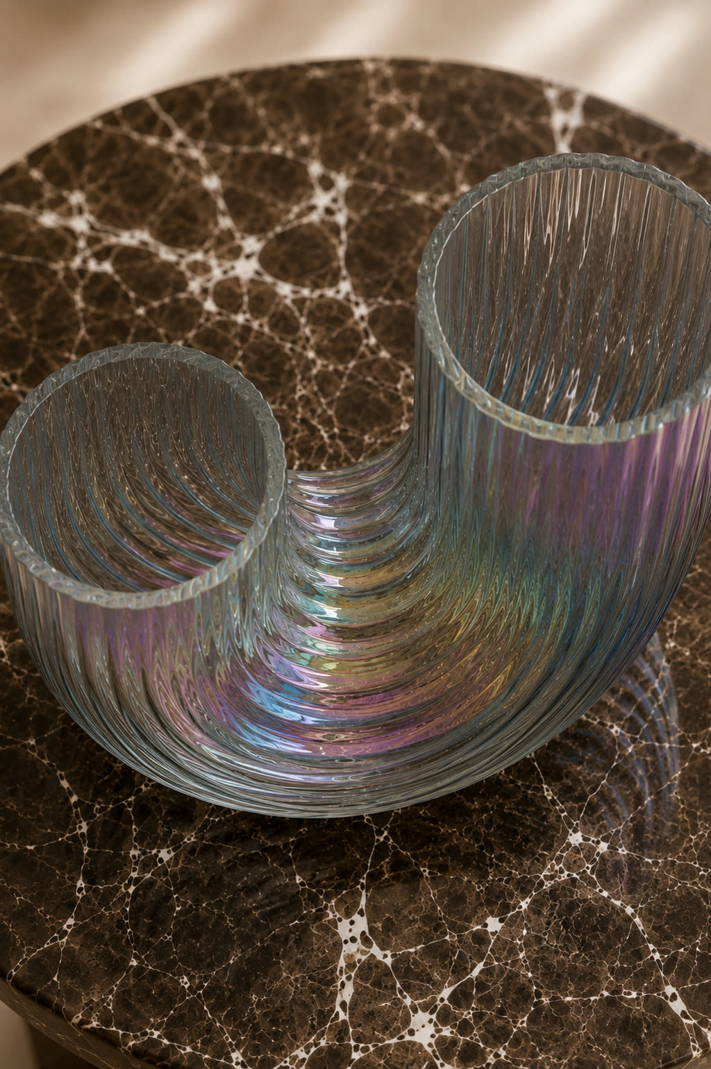 An iridescent glass sculpture, two ribbed tubes joined in a loop, on a dark marble table